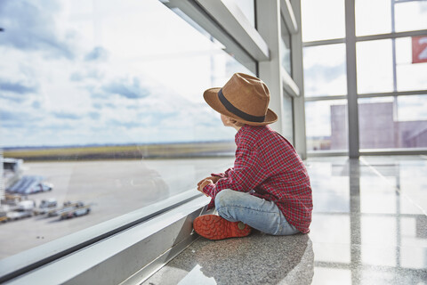 Junge sitzt hinter einer Fensterscheibe auf dem Flughafen und schaut auf das Flugfeld, lizenzfreies Stockfoto