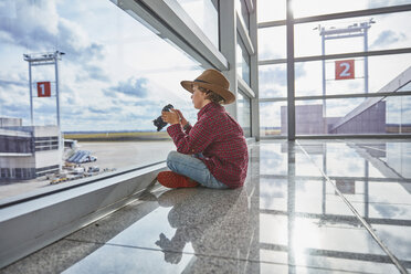 Boy sitting behind windowpane at the airport holding a camera - SSCF00345