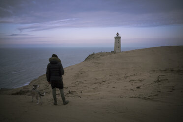 Dänemark, Nordjütland, Rückenansicht eines Mannes mit Hund mit Blick auf den Leuchtturm Rubjerg Knude zur blauen Stunde - REAF00489