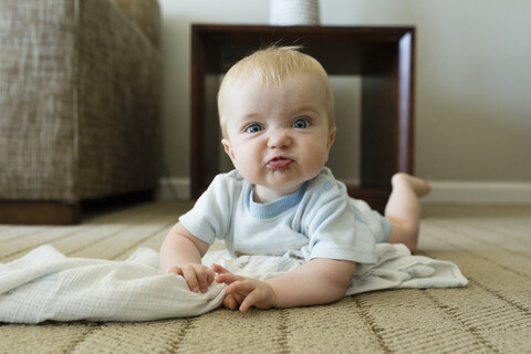 Porträt des niedlichen verspielten kleinen Jungen, der sein Gesicht macht, während er auf dem Teppich zu Hause liegt, lizenzfreies Stockfoto