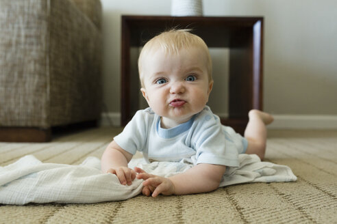Portrait of cute playful baby boy making face while lying on carpet at home - CAVF59210