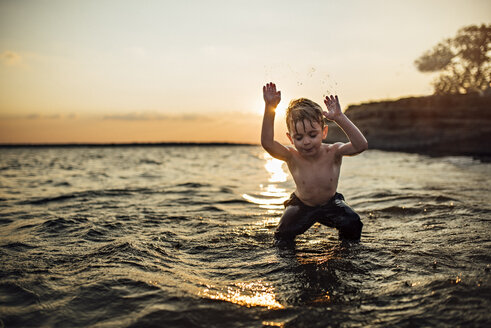 Sorgloser Junge ohne Hemd spielt im Fluss gegen den Himmel bei Sonnenuntergang - CAVF59215