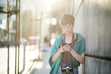 Teenage girl using smart phone while leaning on wall in city - CAVF59224