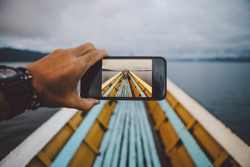 Ausgeschnittene Hand eines Mannes, der mit seinem Smartphone ein Boot gegen den Himmel fotografiert - CAVF59239