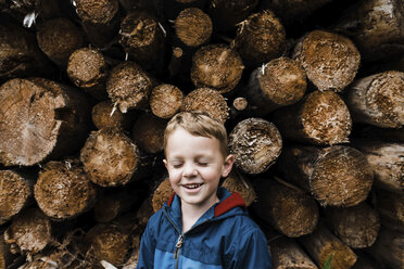 Smiling boy with eyes closed standing by logs - CAVF59267