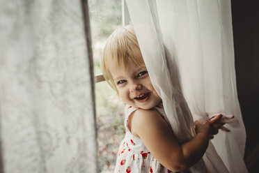 Portrait of cheerful girl playing with curtains by window at home - CAVF59303