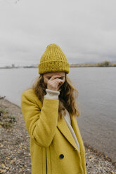 Frau mit Hand im Gesicht, die einen Wintermantel trägt und am Seeufer steht - CAVF59396