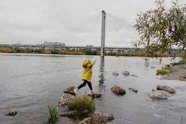 Full length of woman jumping on rocks in lake against bridge - CAVF59398