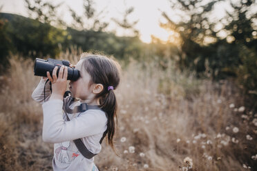 Side view of girl looking through binoculars while standing on field - CAVF59403