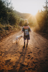 Rückansicht des Vaters, der seine Tochter auf den Schultern trägt, während er mit seinem Hund auf einem unbefestigten Weg an einem sonnigen Tag spazieren geht - CAVF59405