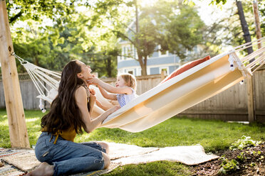 Happy mother playing with daughter sitting in hammock at yard - CAVF59475