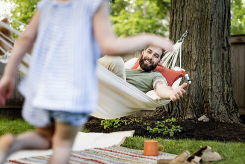 Niedriger Ausschnitt einer Tochter, die auf ihren Vater zuläuft, der sich in einer Hängematte im Garten entspannt - CAVF59477