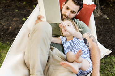 Hoher Blickwinkel der Tochter mit dem Vater, der ein Buch liest, während er in der Hängematte im Garten liegt - CAVF59478