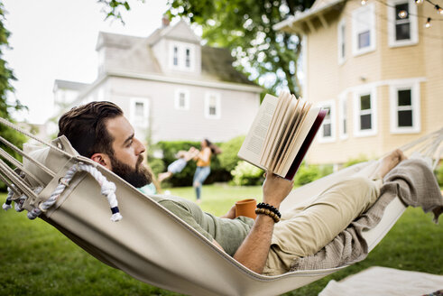 Vater liest ein Buch, während Mutter und Tochter im Hintergrund im Garten spielen - CAVF59480