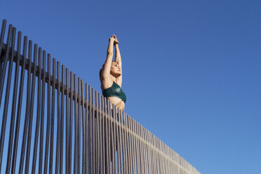Low angle view of young woman stretching arms while exercising on bridge against clear sky - CAVF59559