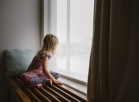 Seitenansicht eines Mädchens, das durch das Fenster schaut, während es zu Hause sitzt, lizenzfreies Stockfoto
