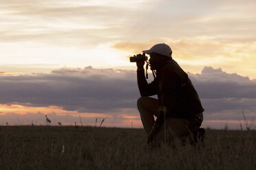 Seitenansicht eines Mannes, der ein Fernglas benutzt, während er auf einem Feld im Serengeti-Nationalpark bei Sonnenuntergang kniet - CAVF59609