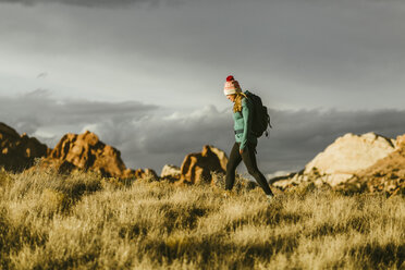 Seitenansicht einer Wanderin mit Rucksack, die auf einer Wiese gegen Felsformationen und einen bewölkten Himmel bei Sonnenuntergang läuft - CAVF59644