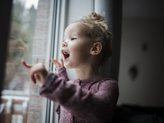 Mädchen mit offenem Mund, das durch das Fenster schaut, während es zu Hause steht - CAVF59654