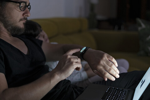 Vater mit Tochter auf der Couch sitzend mit Laptop und Smartwatch in der Nacht, lizenzfreies Stockfoto