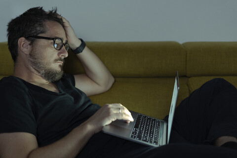 Man lying on couch using laptop at night stock photo