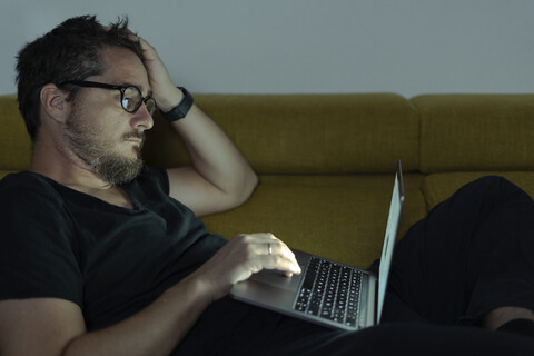 Mann liegt auf der Couch und benutzt nachts einen Laptop, lizenzfreies Stockfoto