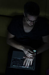 Man sitting on couch using laptop and smartwatch at night - ERRF00299
