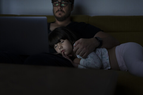 Vater benutzt Laptop nachts auf der Couch, während seine Tochter schläft, lizenzfreies Stockfoto