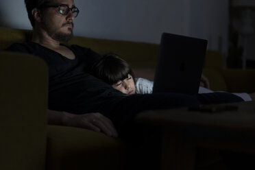 Father using laptop on couch at night with daughter sleeping - ERRF00303