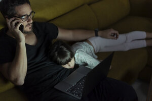 Father talking on cell phone on couch at night with daughter lying next to him - ERRF00306