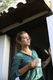 Junge Frau mit Tasse Kaffee steht am Hauseingang und schaut in die Ferne - ERRF00326