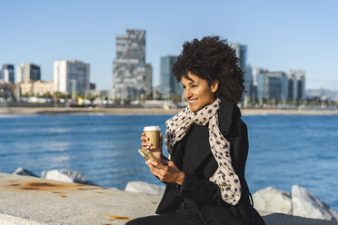 Spain, Barcelona, portrait of smiling with coffee to go and smartphone sitting on a wall - AFVF02068