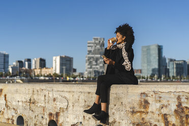 Spanien, Barcelona, Frau mit Smartphone sitzt an einer Wand und trinkt Kaffee zum Mitnehmen - AFVF02069