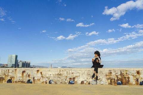 Spanien, Barcelona, Frau mit Smartphone und Kaffee zum Mitnehmen auf einer Mauer sitzend, lizenzfreies Stockfoto
