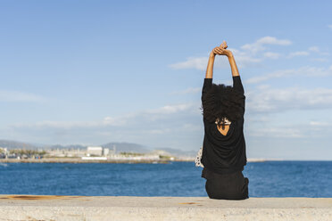 Spanien, Barcelona, Rückenansicht einer schwarz gekleideten Frau, die auf einer Mauer sitzt und auf das Meer schaut - AFVF02077