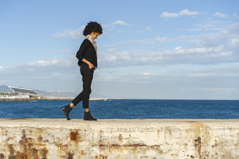 Spanien, Barcelona, lachende, schwarz gekleidete Frau, die im Sonnenlicht auf einer Mauer läuft, lizenzfreies Stockfoto