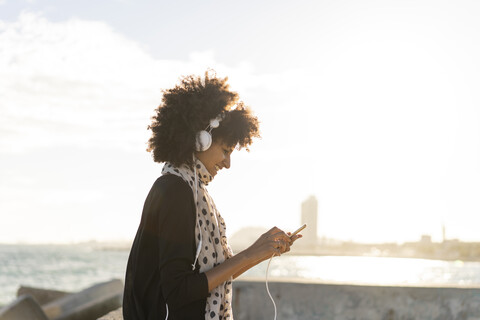 Lächelnde Frau hört Musik mit Smartphone und Kopfhörer in der Nähe des Meeres, lizenzfreies Stockfoto