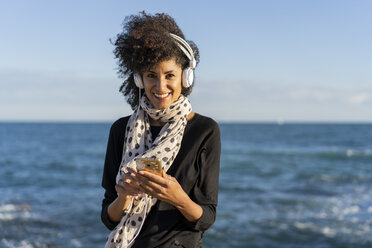 Portrait of smiling woman listening music with smartphone and headphones in front of the sea - AFVF02102