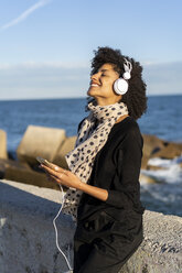 Smiling woman listening music with smartphone and headphones near the sea - AFVF02104