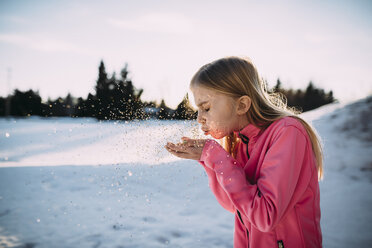 Side view of girl blowing confetti while standing on snow covered field against sky - CAVF59681