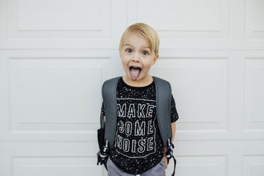 Porträt eines Jungen, der seine Zunge herausstreckt, während er mit einem Rucksack an einer weißen Wand steht - CAVF59697