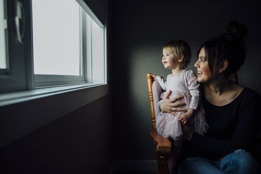 Mutter und Tochter schauen durch das Fenster, während sie auf einem Stuhl zu Hause sitzen - CAVF59707