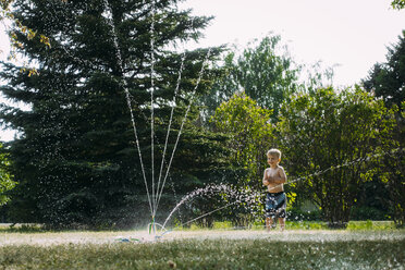 Happy shirtless boy looking at sprinkler spraying water at park - CAVF59722