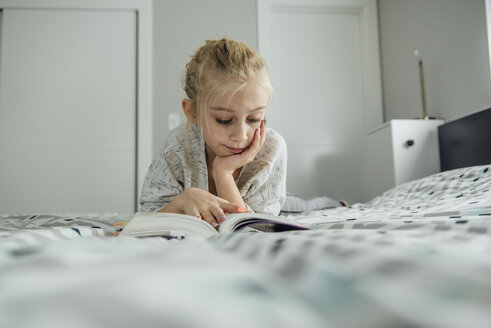 Girl with hand on chin reading book while lying on bed at home - CAVF59743