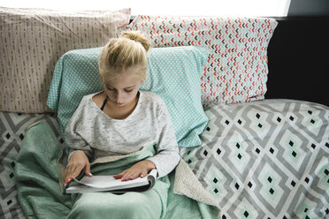 High angle view of girl reading book while leaning on pillows in bedroom - CAVF59749