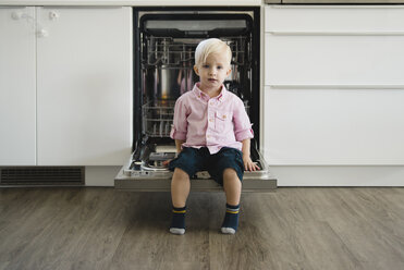 Porträt eines süßen Jungen in voller Länge, der zu Hause auf der Spülmaschinentür sitzt - CAVF59750