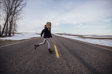 Mädchen läuft in voller Länge auf der Straße inmitten eines schneebedeckten Feldes gegen den Himmel - CAVF59798