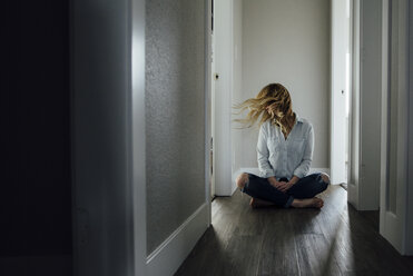 Full length of woman tossing hair while sitting on floor in corridor at home - CAVF59800