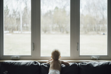 Rückansicht eines kleinen Jungen ohne Hemd, der durch das Fenster schaut, während er zu Hause auf dem Sofa steht - CAVF59804