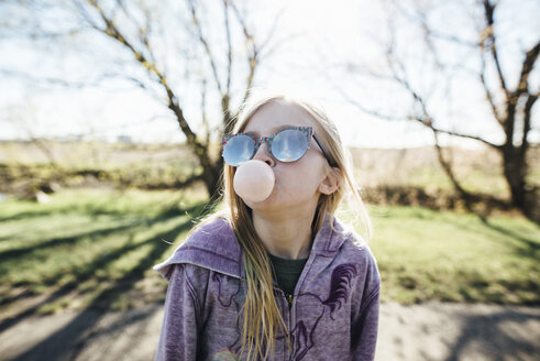 Mädchen mit Sonnenbrille bläst Kaugummi, während sie auf der Straße gegen den Himmel steht - CAVF59820